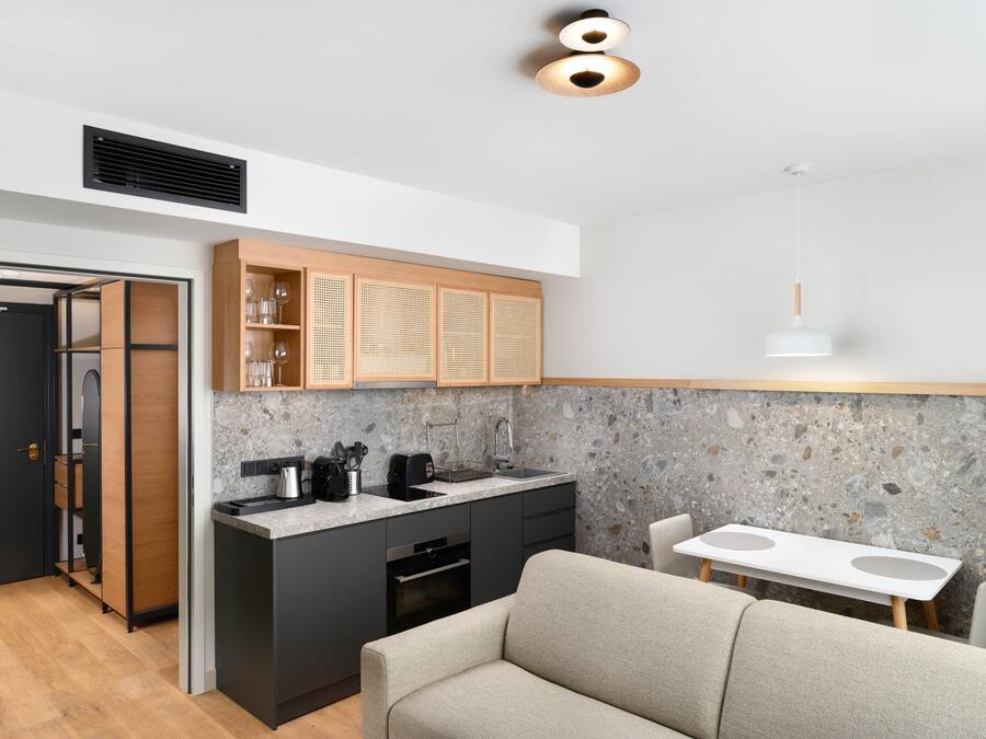 Eine moderne Küche mit eingebautem Herd, schwarzen Schränken und einer Küchenrückwand aus Stein. Im Vordergrund steht ein Esstisch mit weißem Tischlaken und ein beiges Sofa.