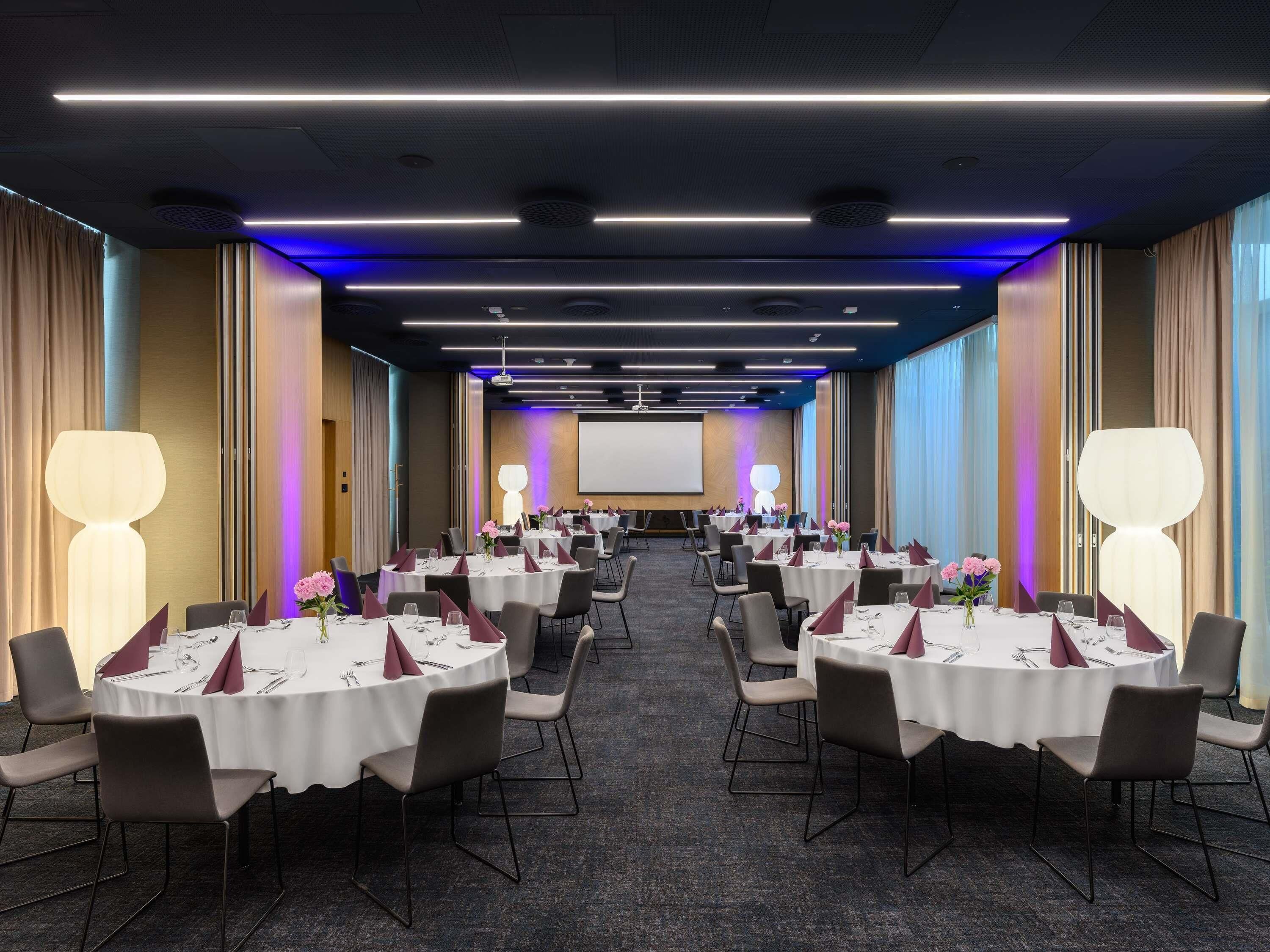 A conference room with multiple tables, chairs, and a large screen at the front.