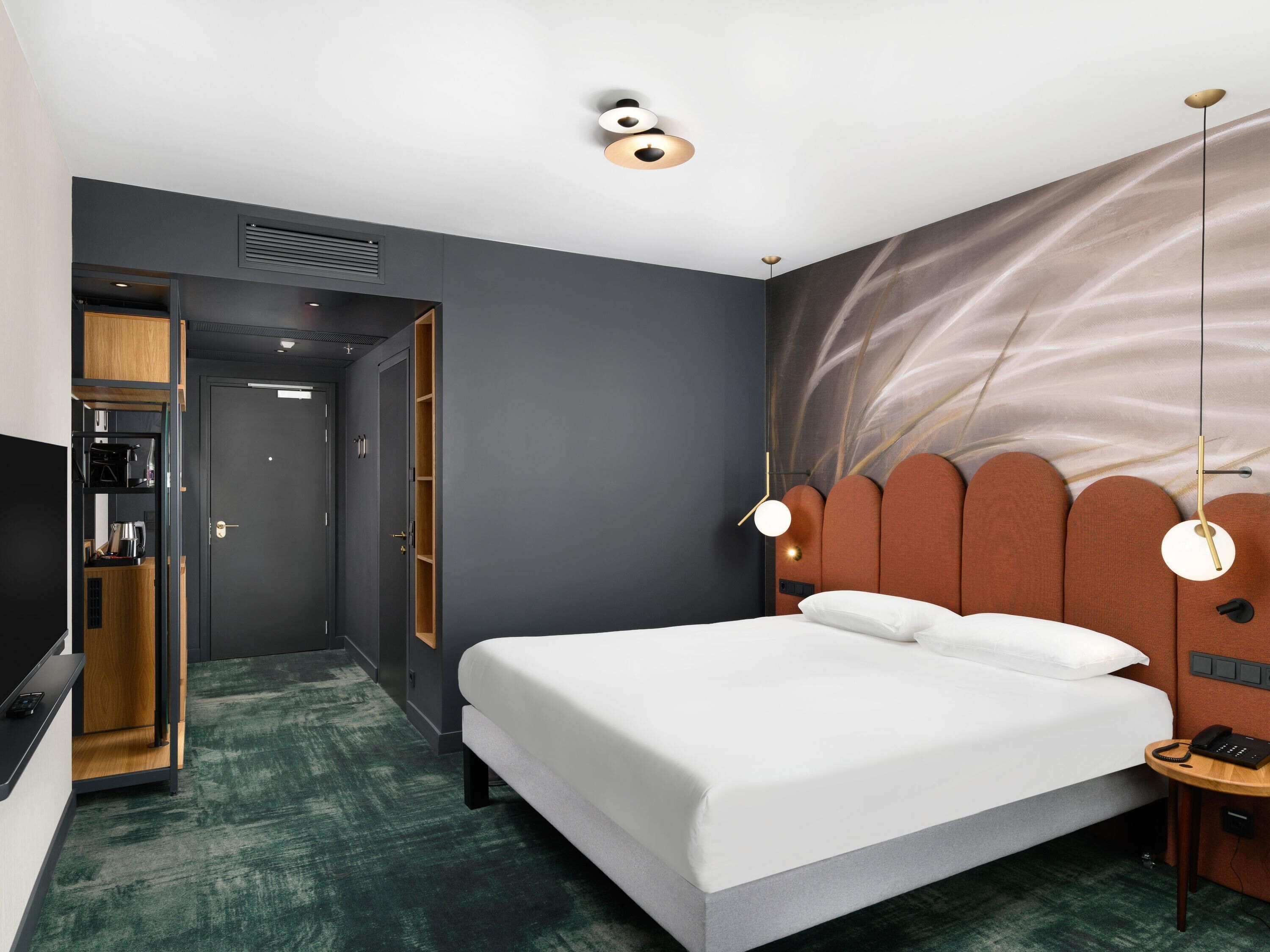 A modern hotel room with a large bed, a desk with a telephone, and a wardrobe.
