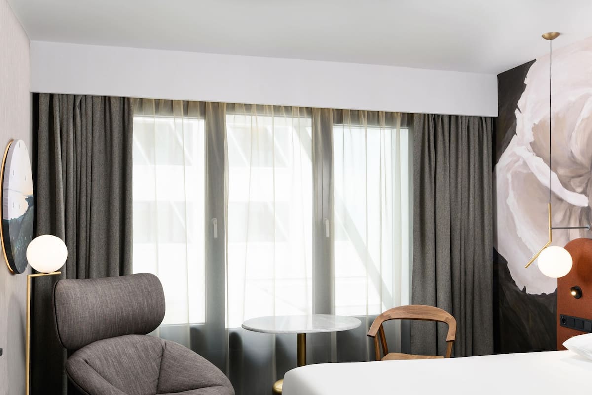 A hotel room with a bed, a chair, a small table, and a large window with curtains.