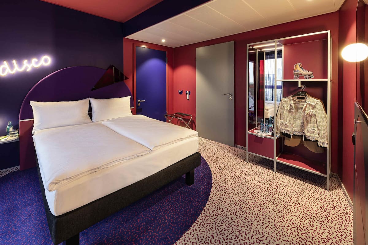 A hotel room with a bed, a wardrobe, a mirror, and a neon sign.