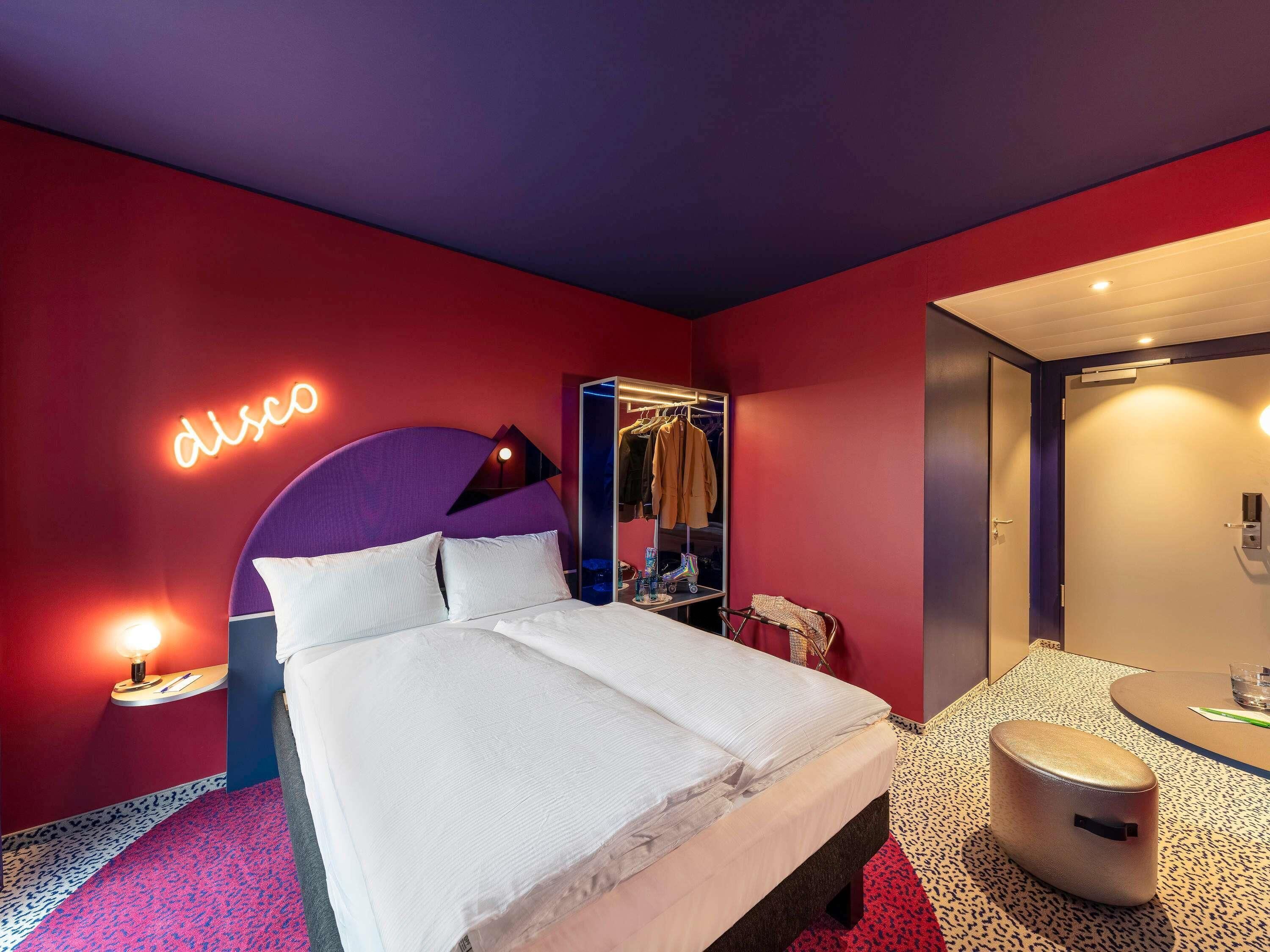 Ett hotellrum med en säng, en garderob, en spegel, en liten pall och en neonreklamskylt.