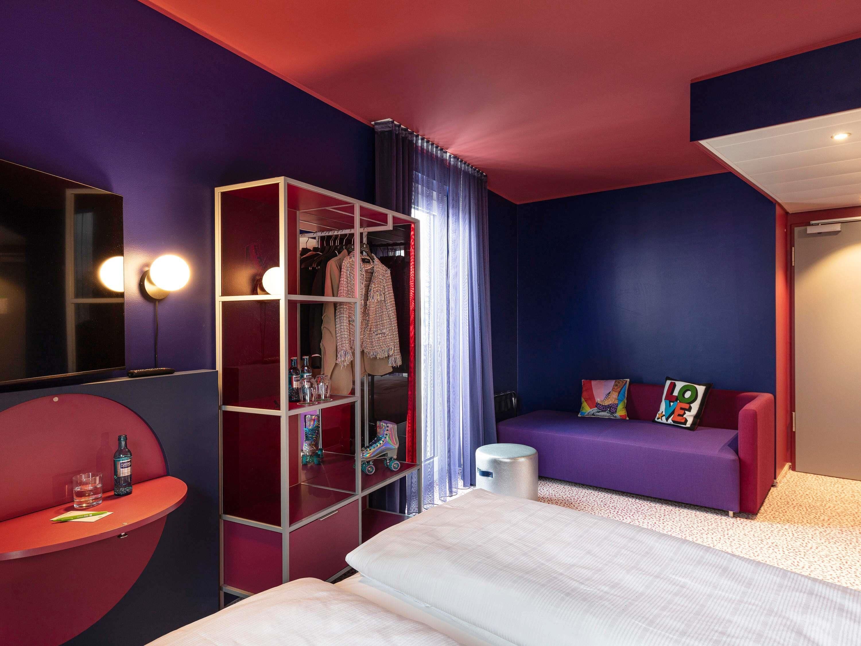 Habitación de hotel moderna con una decoración en tonos morado y rojo, una cama, una zona de estar con sofá, un armario y una mesita con una botella y un vaso.
