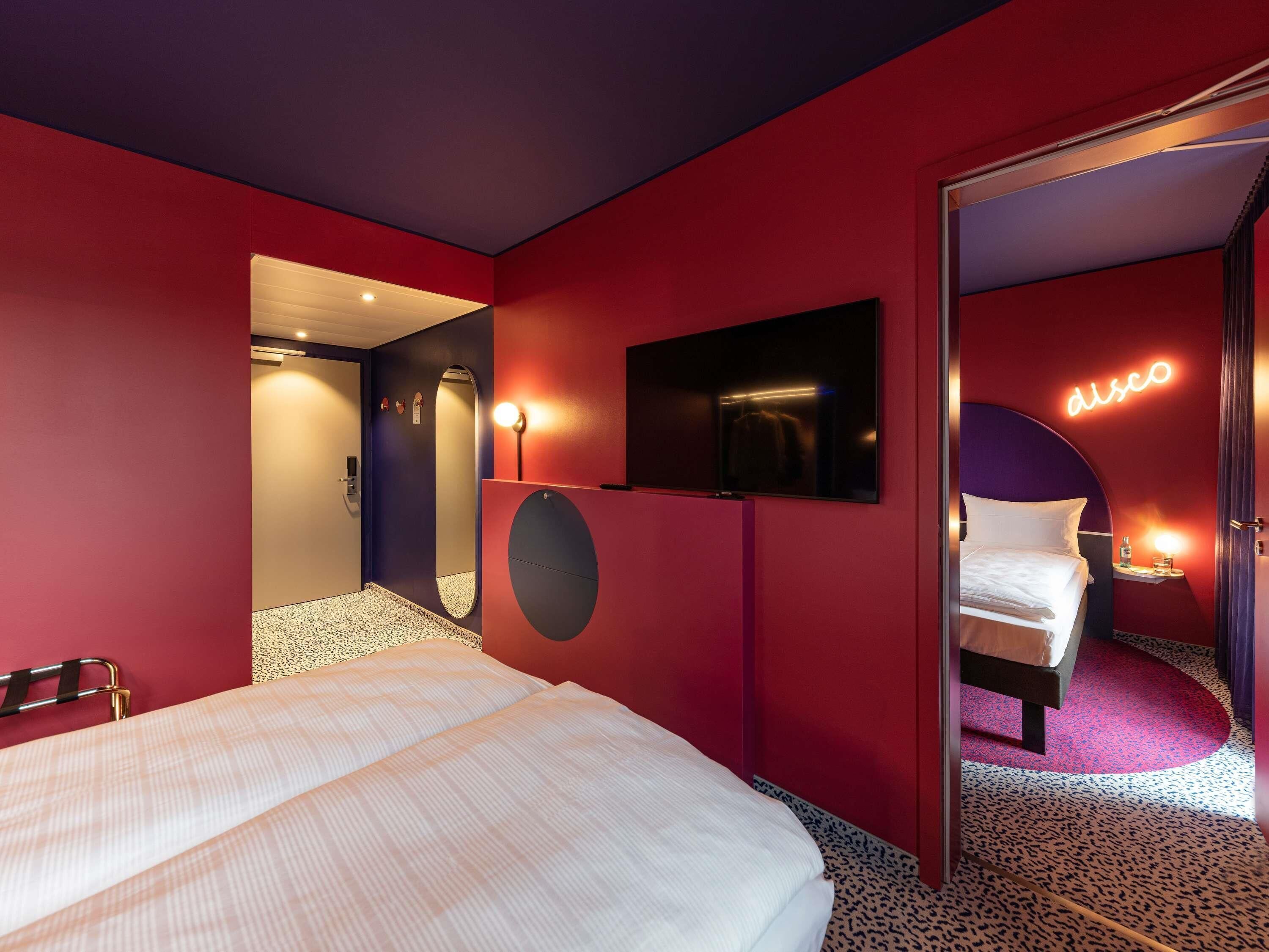 A modern hotel room with a bed, a TV, a mirror, and a neon sign.