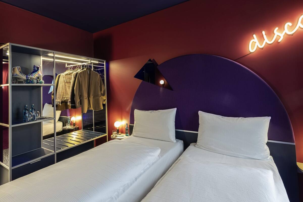A hotel room with two beds, a wardrobe, a mirror, and a neon sign.