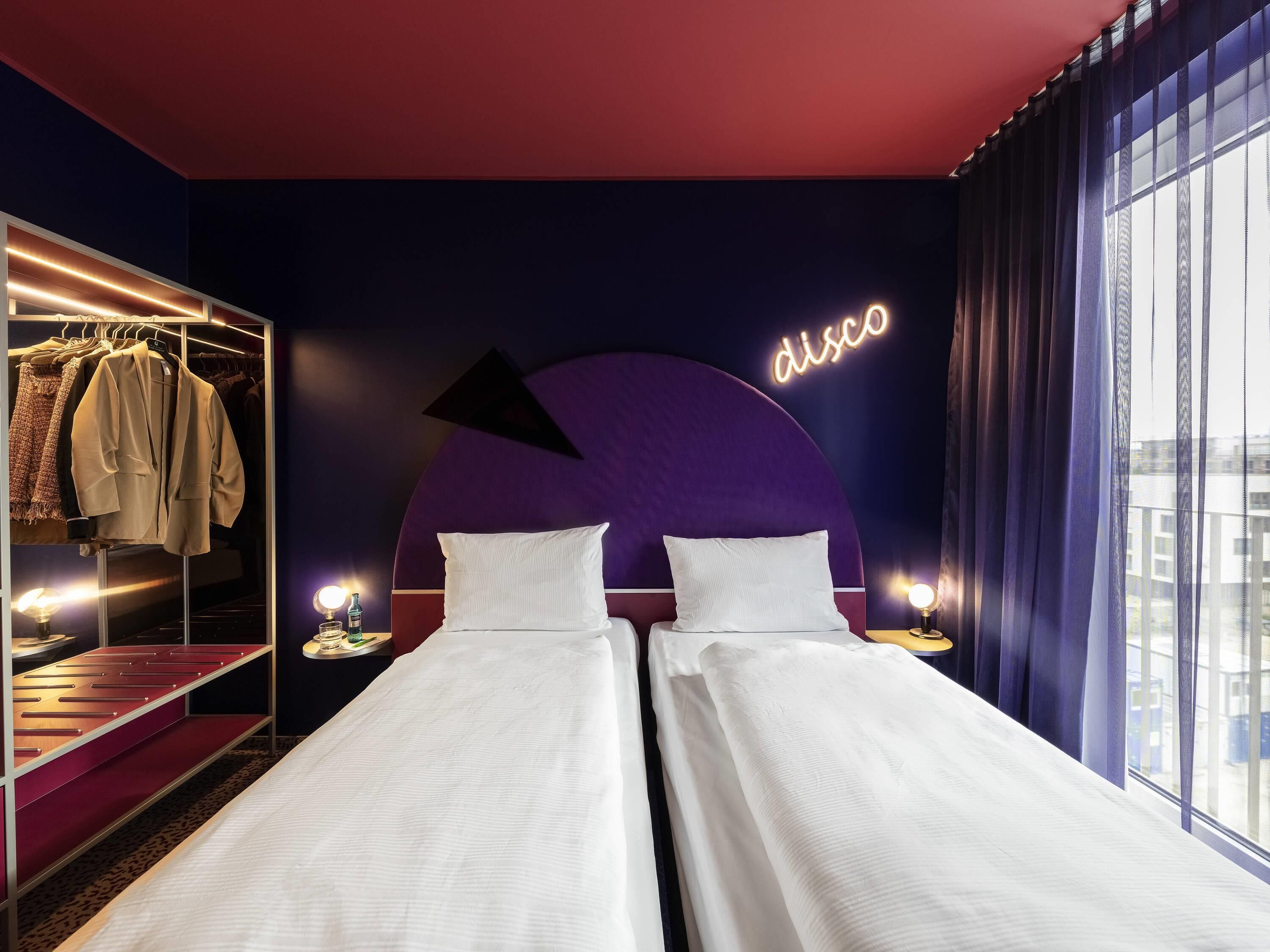 A hotel room with a bed, a closet with clothes, a neon sign, and a window.