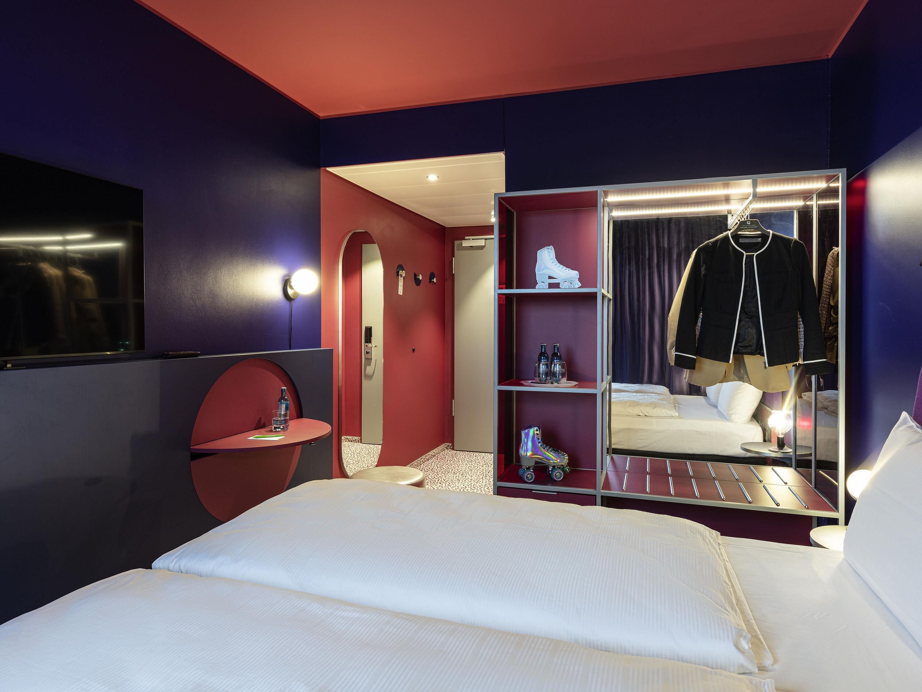 A hotel room with a bed, a wardrobe, a mirror, and a small table with a bottle.