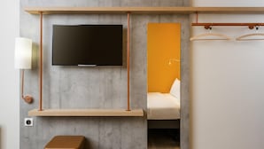 A modern room with a flat-screen TV, a wooden shelf, a mirror, and a hanging light fixture. - Ibis Budget Reims Parc Des Expositions (Reims)
