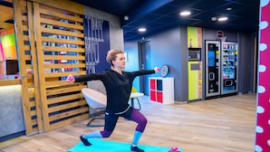 A person stretching on a mat in a room with a wooden slat wall, a vending machine, and a basketball hoop. - Ibis Budget Reims Parc Des Expositions (Reims)