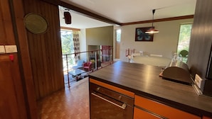 A modern kitchen with wooden cabinets, a stainless steel sink, and a dining area with a red chair and a glass table. - Chalet les Digitales - Rocher du cerf (Le Lioran)