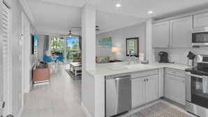 A modern kitchen with white cabinets, a dishwasher, and a stove. There is a dining area with a table and chairs, and a view of palm trees through the window.