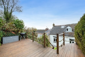 A wooden deck with a barbecue grill, surrounded by greenery and overlooking a residential area with houses.