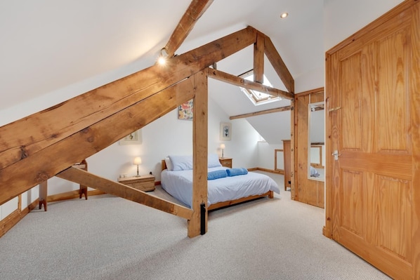 A bedroom with a sloped ceiling, wooden beams, a bed, a nightstand, a lamp, and a wardrobe.