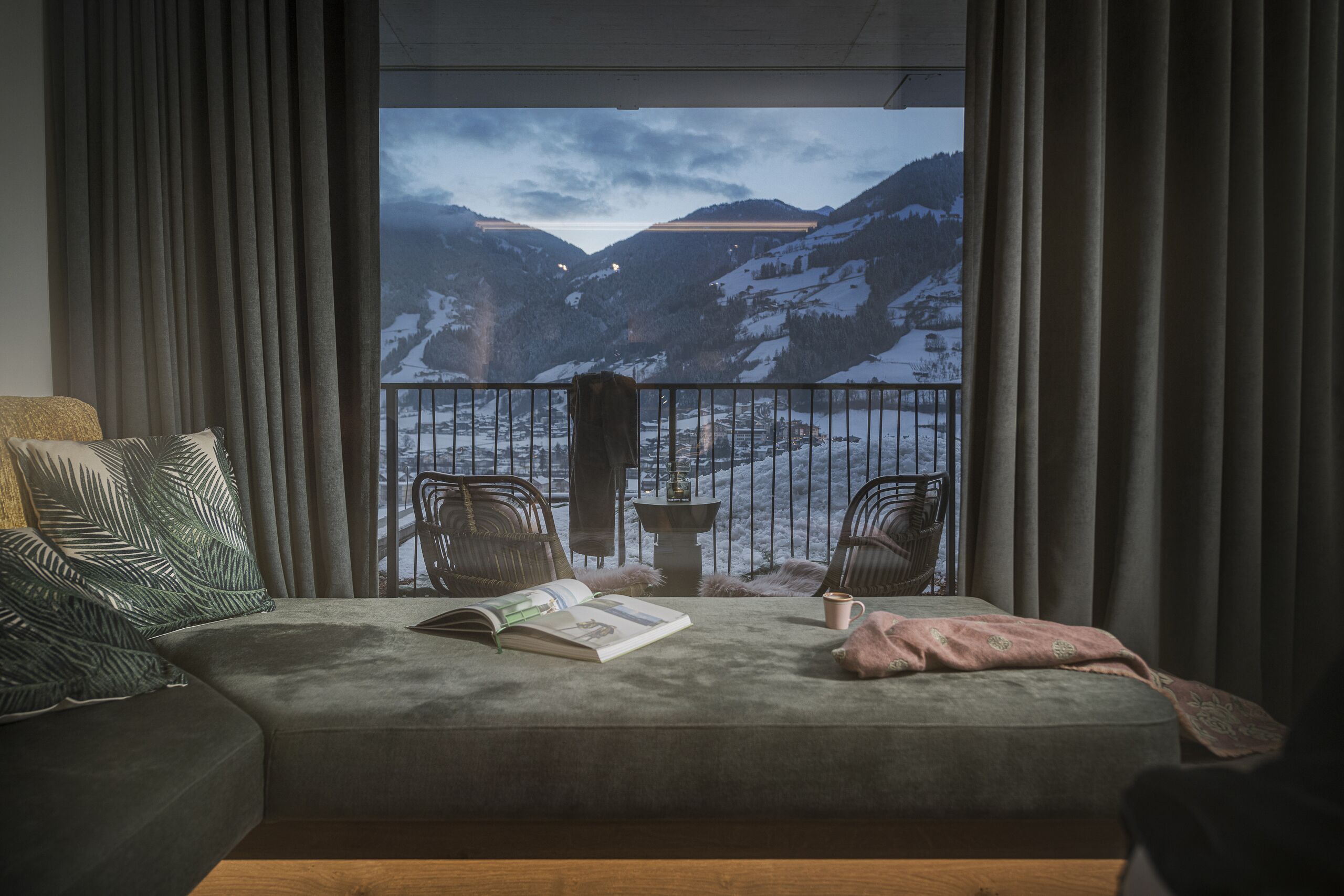 A cozy room with a grey sofa, a book, and a mug, overlooking a snowy mountain landscape through a balcony with a metal railing.