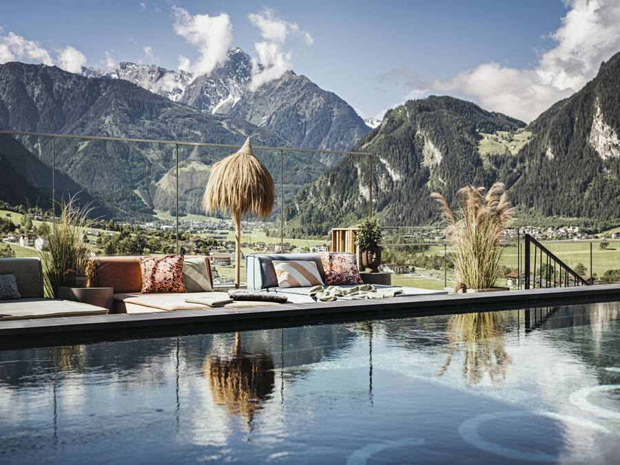 Un espace piscine offrant une vue sur les montagnes, une structure dotée d’un toit de chaume et des sièges d’extérieur agrémentés de coussins.