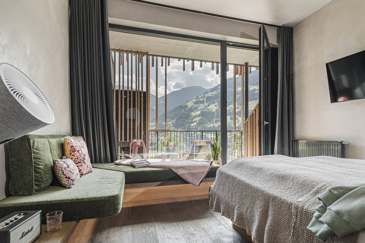 Ein Schlafzimmer mit großem Fenster, einem Bett, einer Couch, einem Heizkörper und einem Fernseher an der Wand.