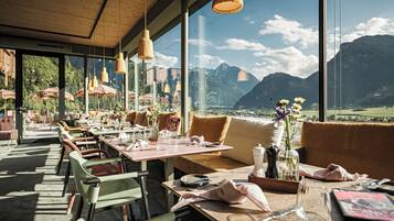 Un restaurante con mesas dispuestas para comer, ventanas amplias con vistas a la montaña y lámparas colgantes.