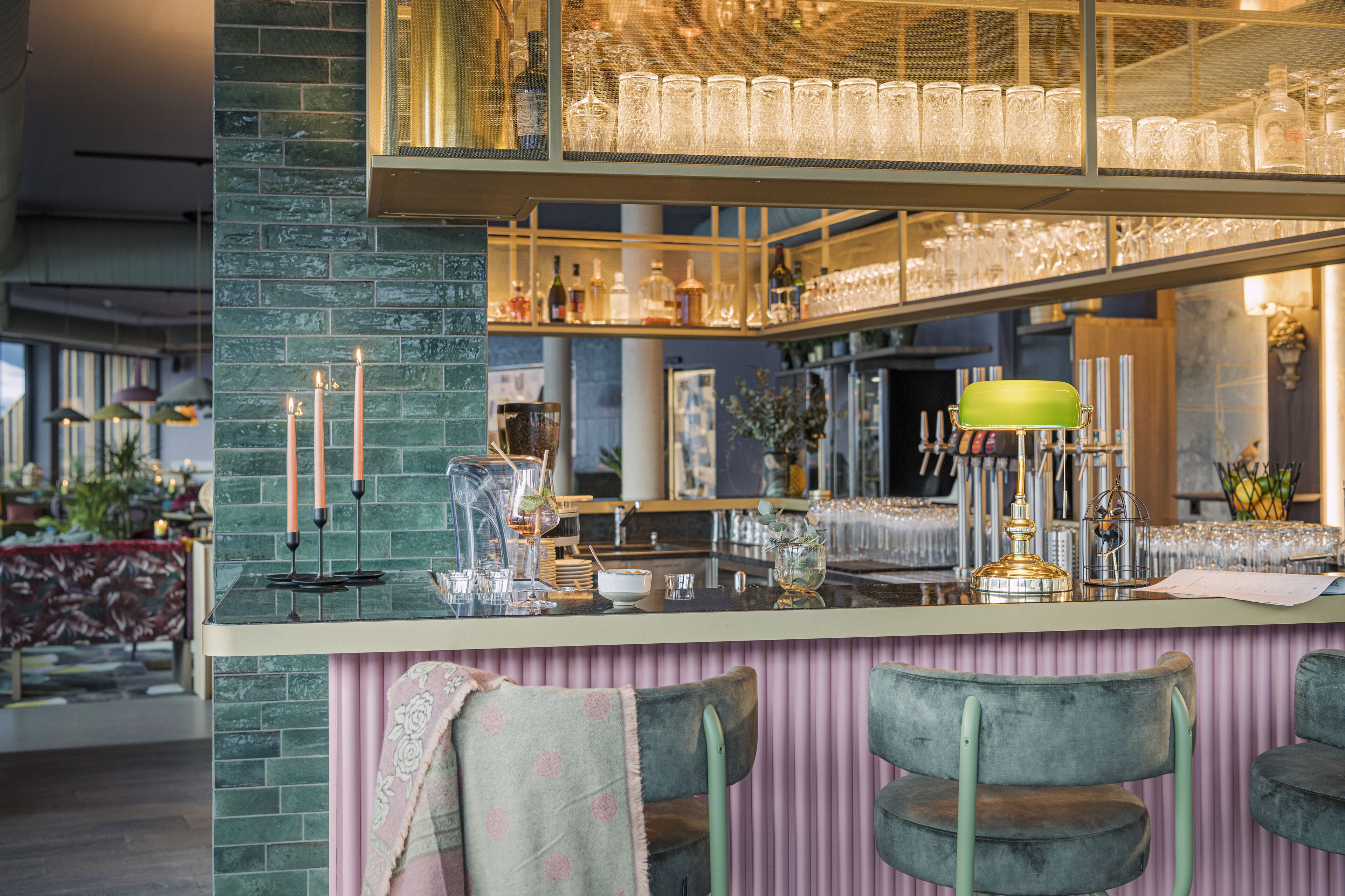 A bar area with a green-tiled counter, bar stools, and a mirrored wall displaying glasses and bottles.