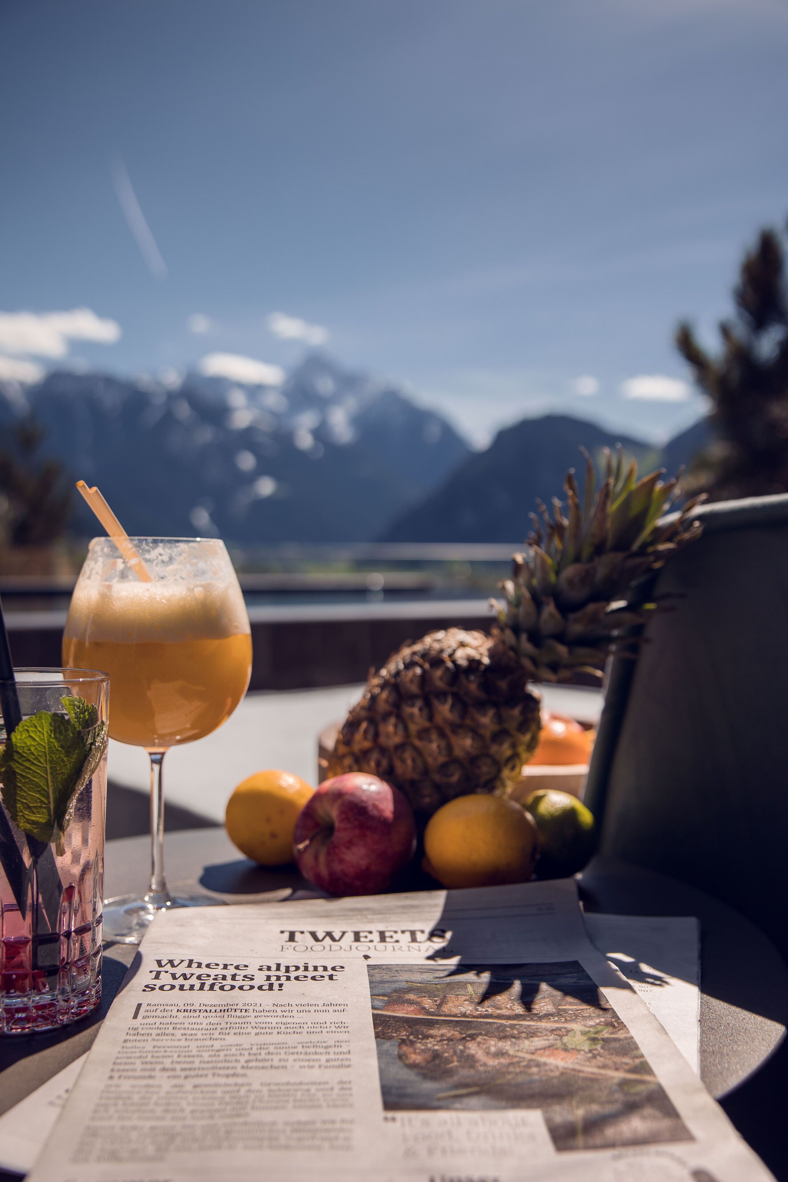 Ein Tisch mit einem Getränk, Früchten und einer Zeitung. Im Hintergrund sind Berge zu sehen.