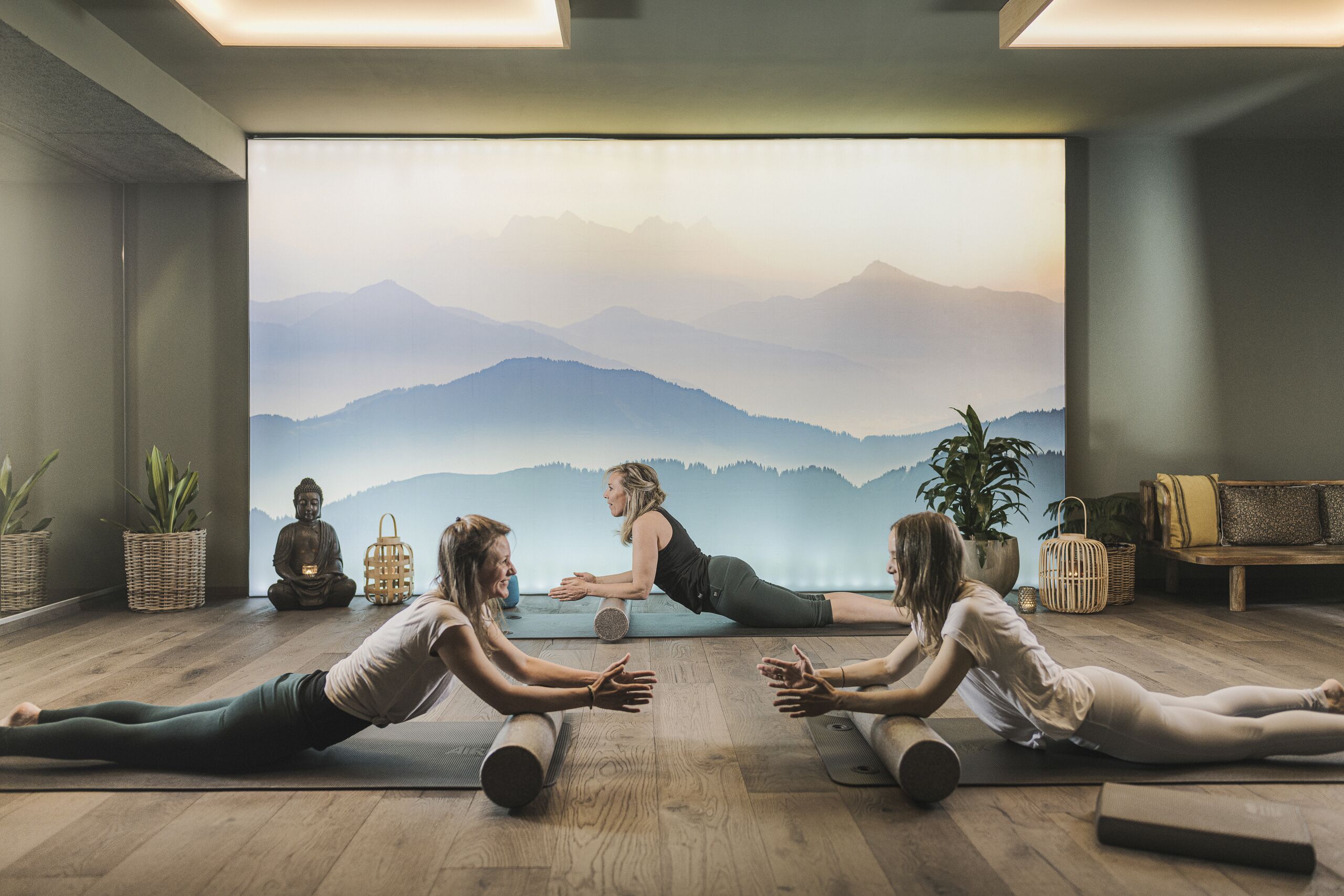 Una clase de yoga en una habitación con piso de madera, un gran mural de montañas y una estatua de Buda.