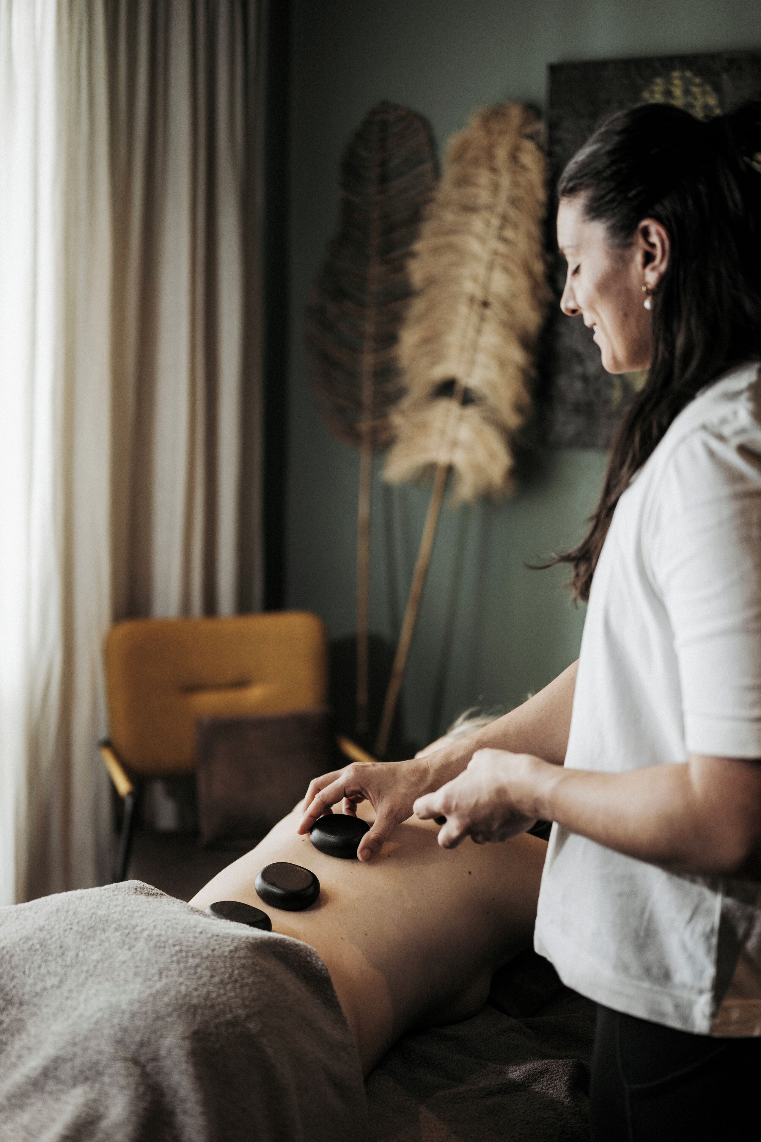 Eine Massageanwendung in einem Raum mit einer großen Pflanze, einem Sessel und einem fenster mit Vorhängen.