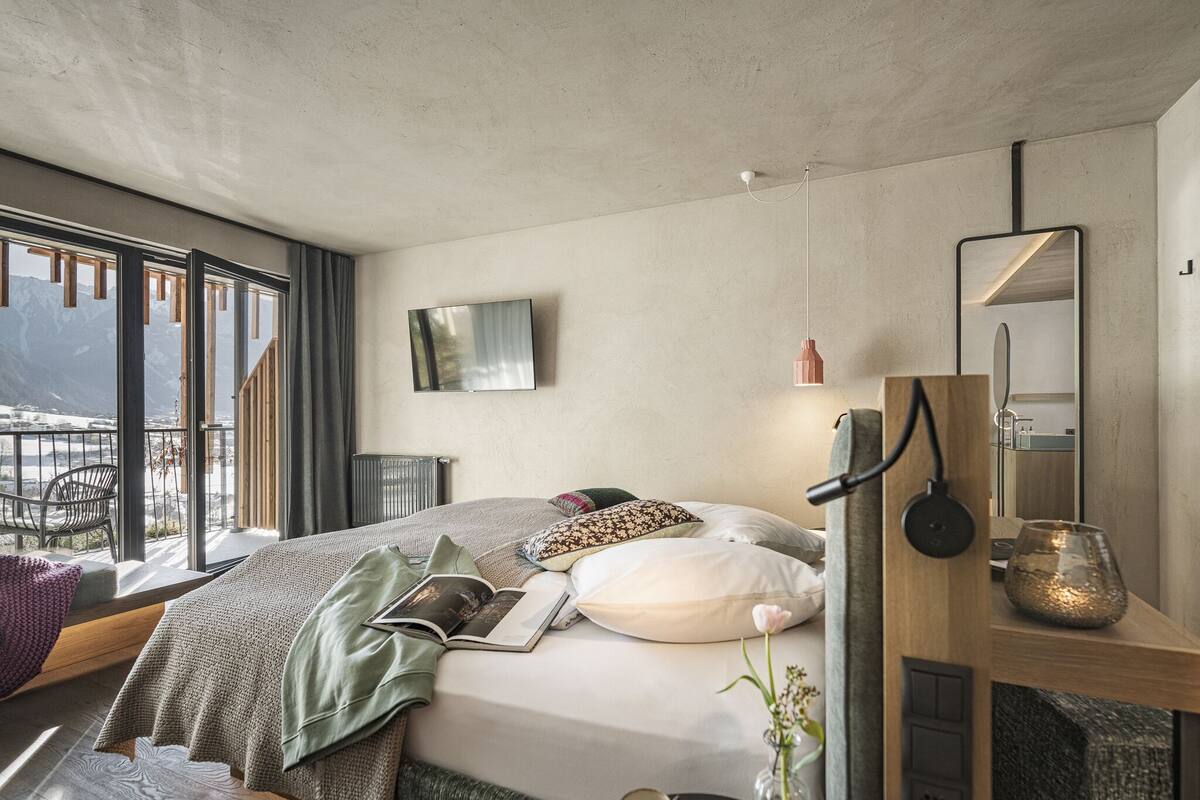 Ein modernes Schlafzimmer mit einem großen Bett, einem Wand-Fernseher, einem Nachttisch mit Lampe und Blick auf Berge durch große Fenster.