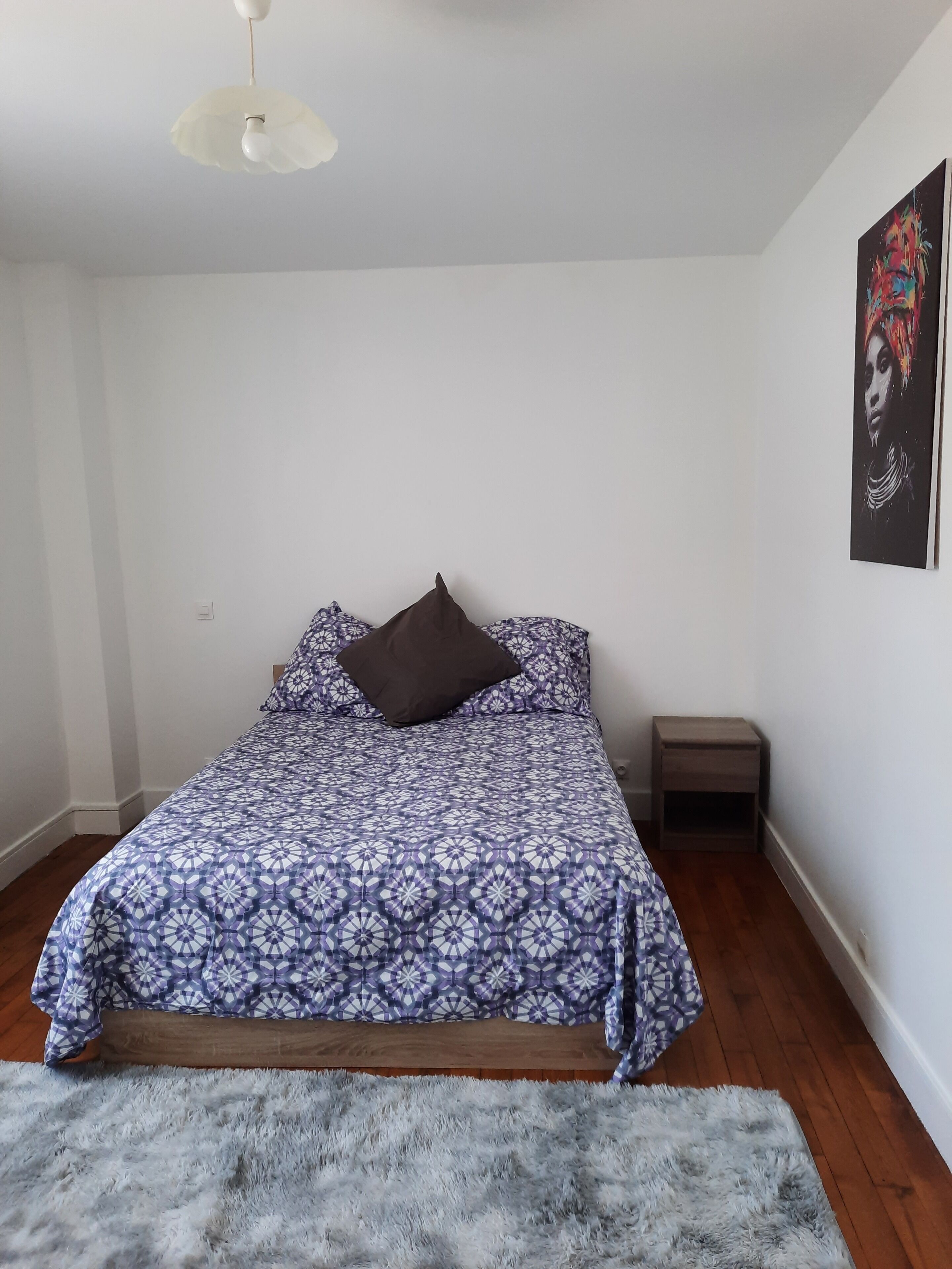 Une chambre à coucher avec une couvre-lit à motifs, un tapis moelleux, une petite table de chevet et un luminaire au plafond.