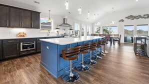 A modern kitchen with dark cabinets, a large island, bar stools, and a view of the outdoors. - All Inn and Then Some: Pool, Spa, Theater Room (Corolla)