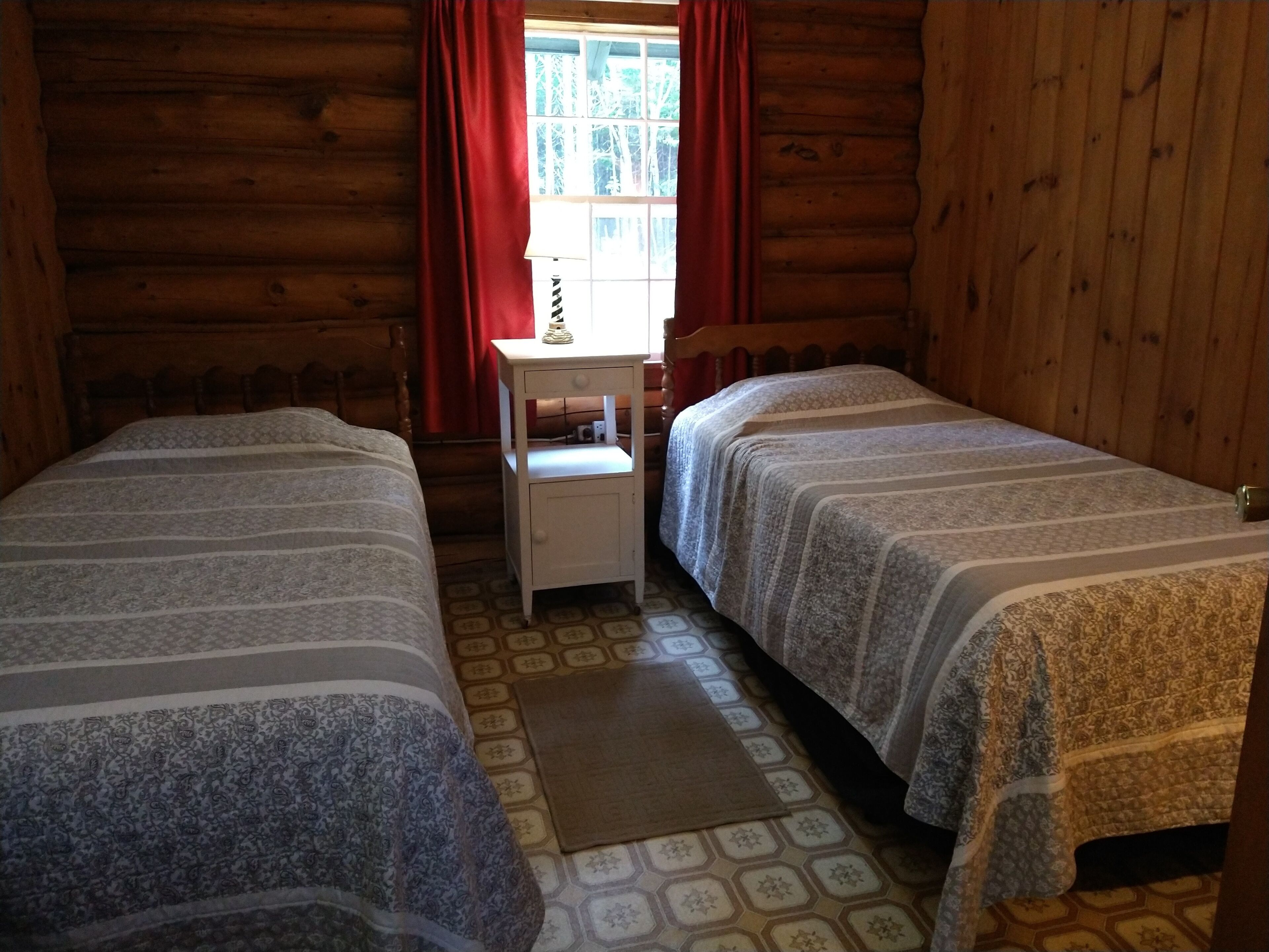 A log cabin room with two beds, a small table, and a window with red curtains.