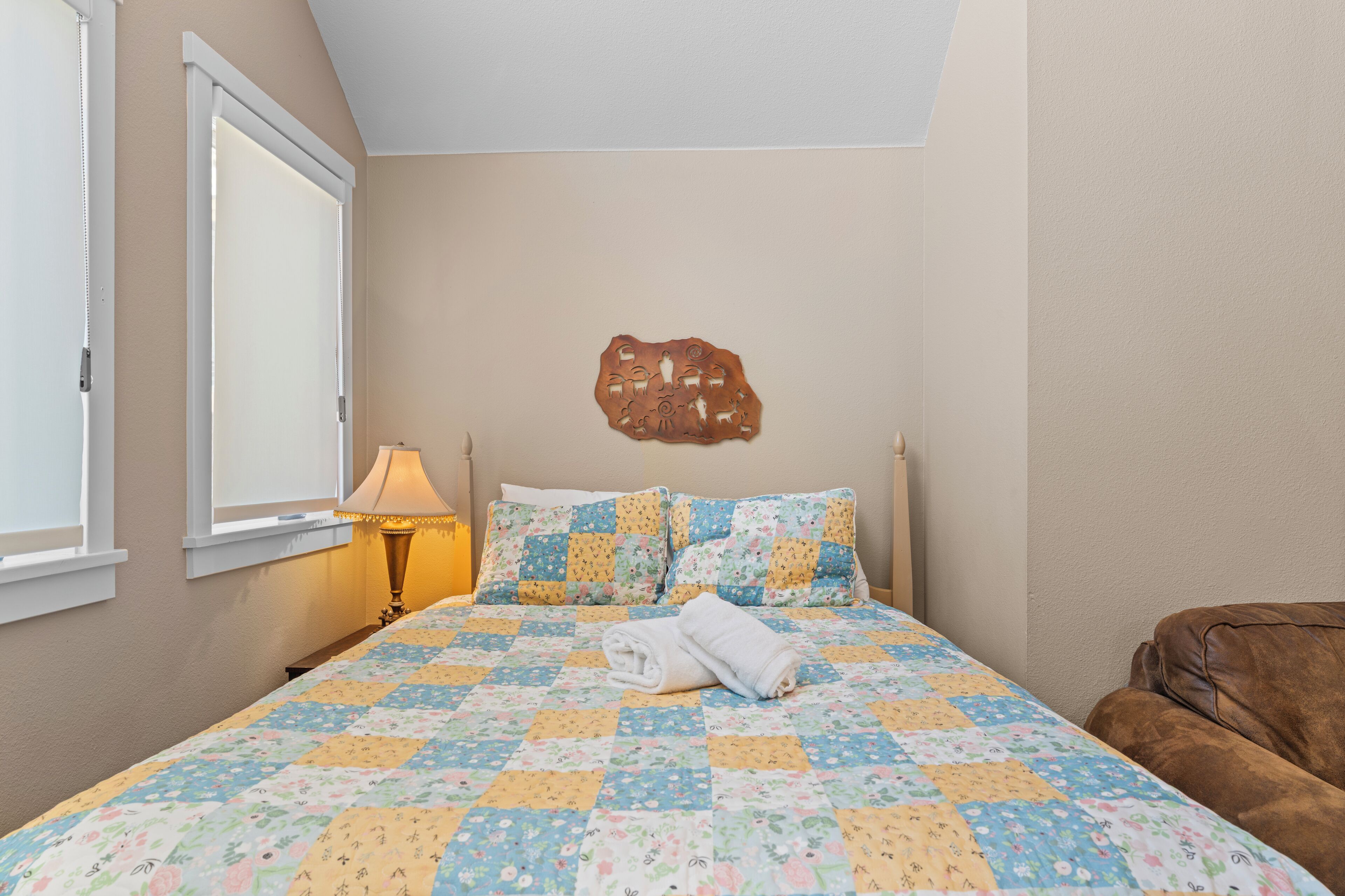A bedroom with a quilted bedspread, a decorative wall piece, a bedside lamp, and a brown leather chair.