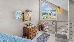 Ein Schlafzimmer mit großem Fenster, einer Holzkommode, einem Spiegel und einem kleinen Tisch mit Lampe.