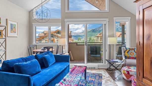 Ein geräumiges Wohnzimmer mit einem blauen Sofa, einem farbenfrohen Teppich und Blick auf eine Berglandschaft durch große Fenster.