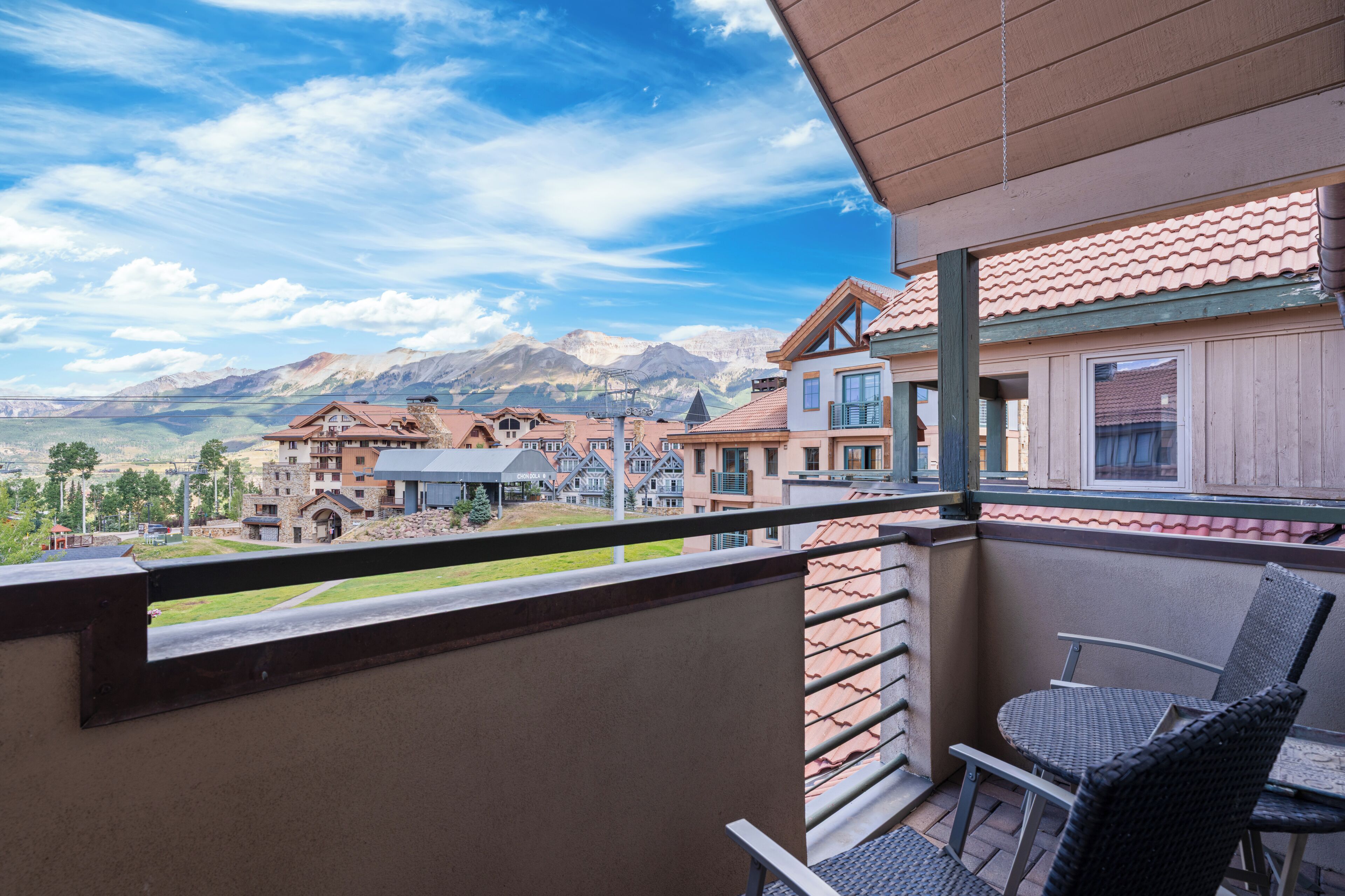 Ein Balkon mit Korbstühlen, Blick auf Bergdörfer und einen klaren Himmel.