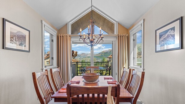 A dining area with a wooden table and chairs, a chandelier, and a view of a mountainous landscape through a glass door. - Blue Mesa Lodge 40B by AvantStay | Ski In/ Ski Out Unit w/ Balcony! (Telluride)