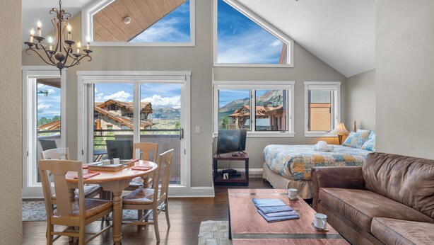 A spacious living room with a high ceiling, large windows, a wooden dining table, and a comfortable sofa.