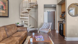 A spacious living area with a brown leather sofa, a wooden coffee table with magazines, a grey armchair, and a staircase leading to an upper level.