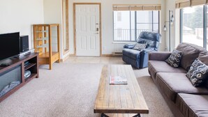 Sala de estar con televisor de pantalla plana, mueble de madera para entretenimiento, mesa de centro, sofá y ventana con persianas.