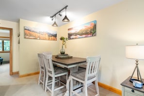 Un comedor con una mesa de madera, cuatro sillas blancas, dos lámparas colgantes fijadas a la pared y dos cuadros de paisajes.