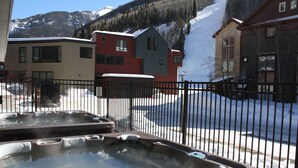 A hot tub area with a metal fence, a building with a red and gray facade, and a snowy mountain backdrop. - Cimarron Lodge 14 by AvantStay | Ski-In/Ski-Out Property w/ Two Hot Tubs! (Telluride)