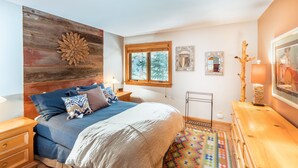 A bedroom with a wooden headboard, a bed with blue bedding, a colorful rug, a dresser, and a window with a view of trees.