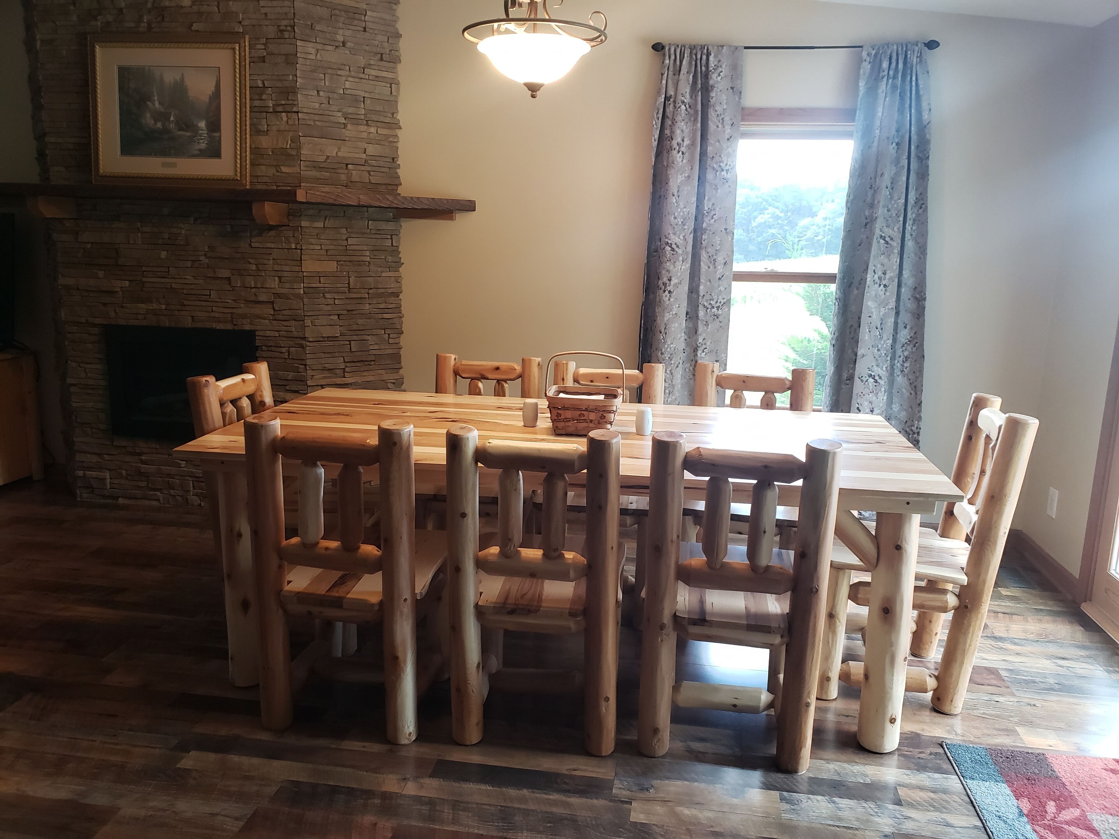 A dining area with a stone fireplace, wooden chairs, a wooden table, and a window with curtains.