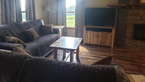 A living room with a sofa, a coffee table, a TV on a wooden stand, and a stone fireplace. - Mountain View Retreat - on 5 private acres only 6 miles from Ohiopyle (Mill Run)