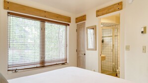 Un dormitorio con una cama grande, cabecera de madera, una ventana con persianas, una puerta que da a un baño y un espejo pequeño en la pared.