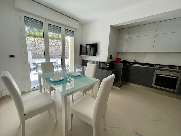 A modern kitchen with a dining area, featuring a glass table, white chairs, and a TV mounted above the counter.