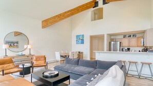 Ein modernes Wohnzimmer mit einer grauen Eckcouch, Ledersesseln, einem Couchtisch und einem Essbereich mit Holztisch und Stühlen. Im Hintergrund ist eine Küche mit Holzschränken und einem Kühlschrank zu sehen.