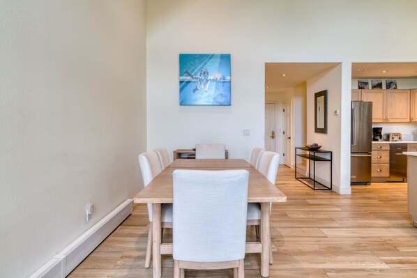 A dining area with a wooden table and chairs, a kitchen in the background, and a painting on the wall.
