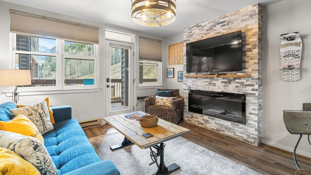 A modern living room with a stone fireplace, a large flat-screen TV, a blue sofa, and a wooden coffee table. - Etta Place 6 by AvantStay | Ski In/ Ski Out Unit! (Telluride)