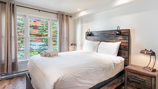Une chambre avec un grand lit, une tête de lit en bois, des lampes de chevet et une vue sur la verdure par la fenêtre.