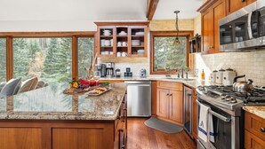 Una cocina con armarios de madera, un refrigerador de acero inoxidable, un horno empotrado y un ventanal con vistas a un bosque.