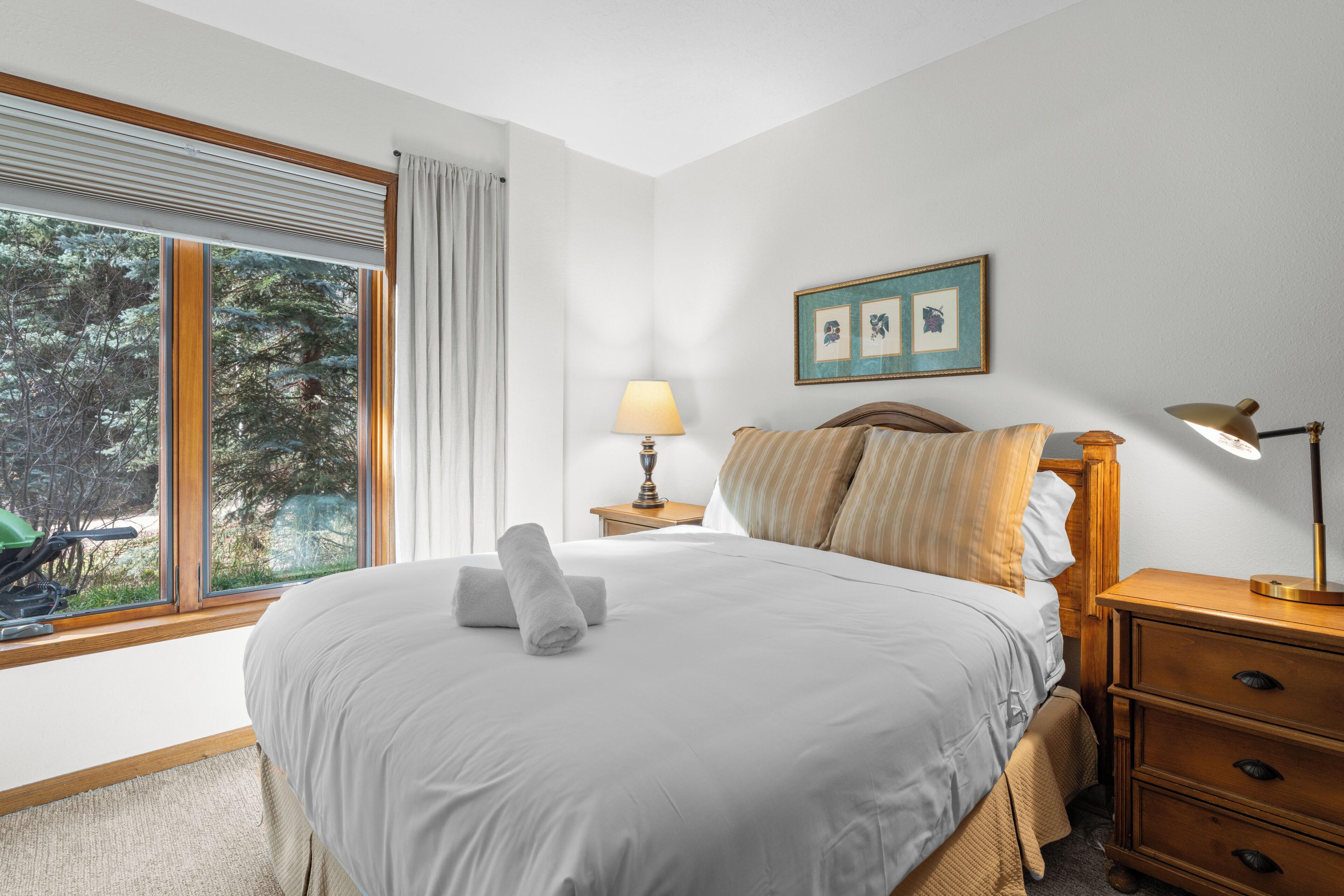 A neatly made bed with a rolled towel, a bedside lamp, a framed picture, and a window with curtains.